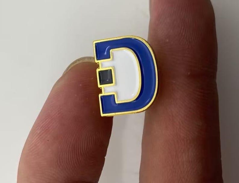 May include: A gold-toned metal lapel pin shaped like the letter D, featuring a bold blue enamel border and a white enamel center. A small, square, dark blue accent is embedded on the left side of the letter, adding a distinct detail to the design.