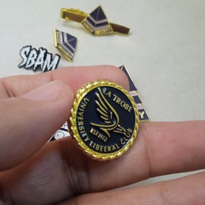 May include: A round gold-toned metal pin with a twisted rope border and a black center featuring a gold wing graphic and the text LA TROBE UNIVERSITY LIBEERAL CLUB EST 1972. Additional metal pins and a tie bar are visible in the background.