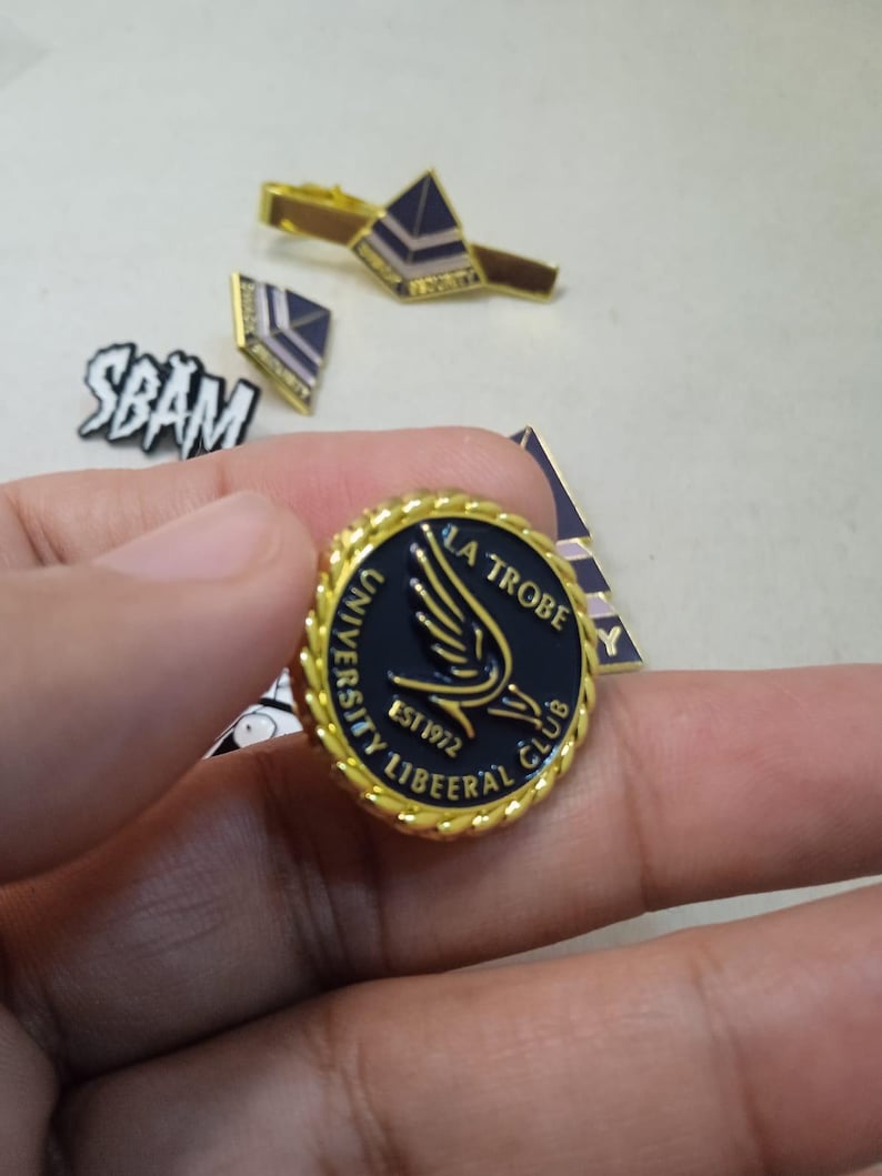May include: A round gold-toned metal pin with a twisted rope border and a black center featuring a gold wing graphic and the text LA TROBE UNIVERSITY LIBEERAL CLUB EST 1972. Additional metal pins and a tie bar are visible in the background.