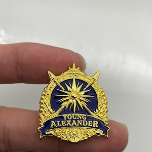May include: A gold and dark blue enamel pin featuring a central starburst design, crossed spears, and decorative laurel leaves. A dark blue banner across the bottom displays the text YOUNG ALEXANDER in gold capital letters with a small circular emblem.