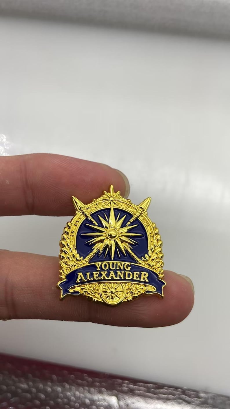 May include: A gold and dark blue enamel pin featuring a central starburst design, crossed spears, and decorative laurel leaves. A dark blue banner across the bottom displays the text YOUNG ALEXANDER in gold capital letters with a small circular emblem.