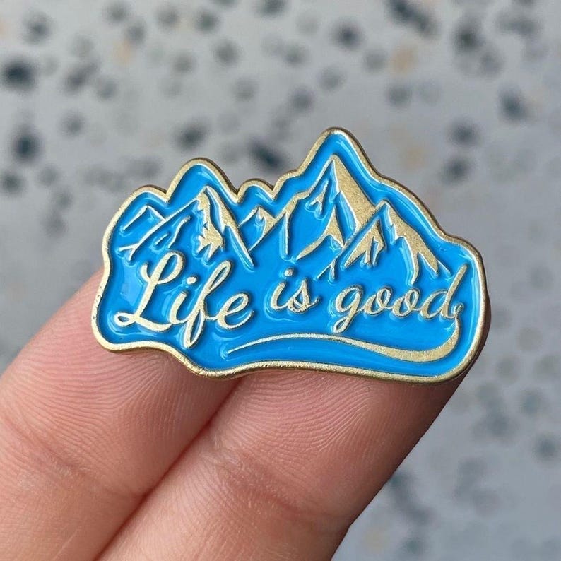 May include: A blue enamel pin with a gold border and the phrase "Life is good." The pin features a mountain range design in gold, set against a vibrant blue background. This accessory is perfect for adding a touch of positivity to any outfit.