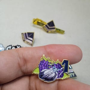 May include: A gold-toned metal enamel pin featuring a cluster of purple grapes with green leaves, a white and grey spaceship, and a dark blue number one. The pin has a polished gold border and a detailed graphic design on the front surface.