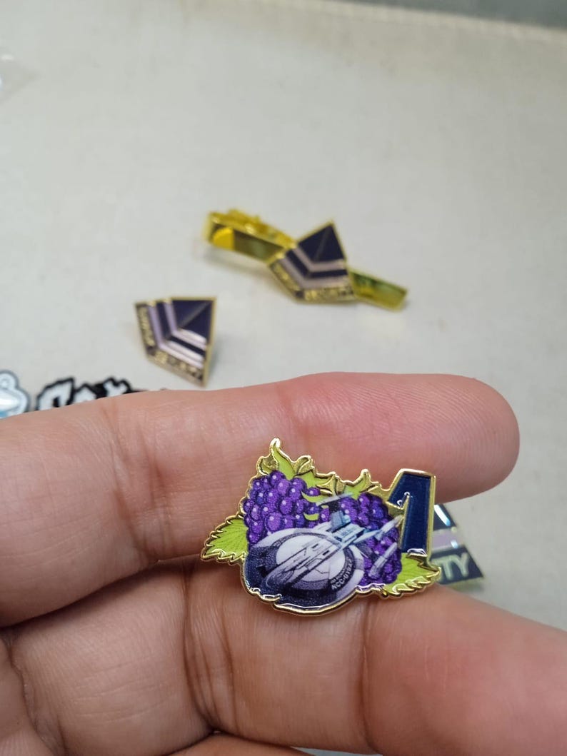 May include: A gold-toned metal enamel pin featuring a cluster of purple grapes with green leaves, a white and grey spaceship, and a dark blue number one. The pin has a polished gold border and a detailed graphic design on the front surface.