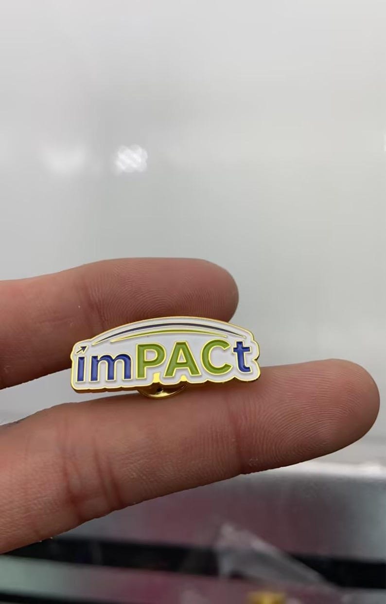 May include: A gold-toned metal lapel pin featuring the word imPACT in a mix of blue and green enamel lettering. The design includes a white background with a thin curved line above the text and a small arrow pointing upward from the letter i.