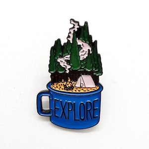 May include: A blue enamel pin shaped like a mug with the word "EXPLORE" on the side. The mug contains a camping scene with a tent, campfire, and trees. The trees are green, and the smoke is white.