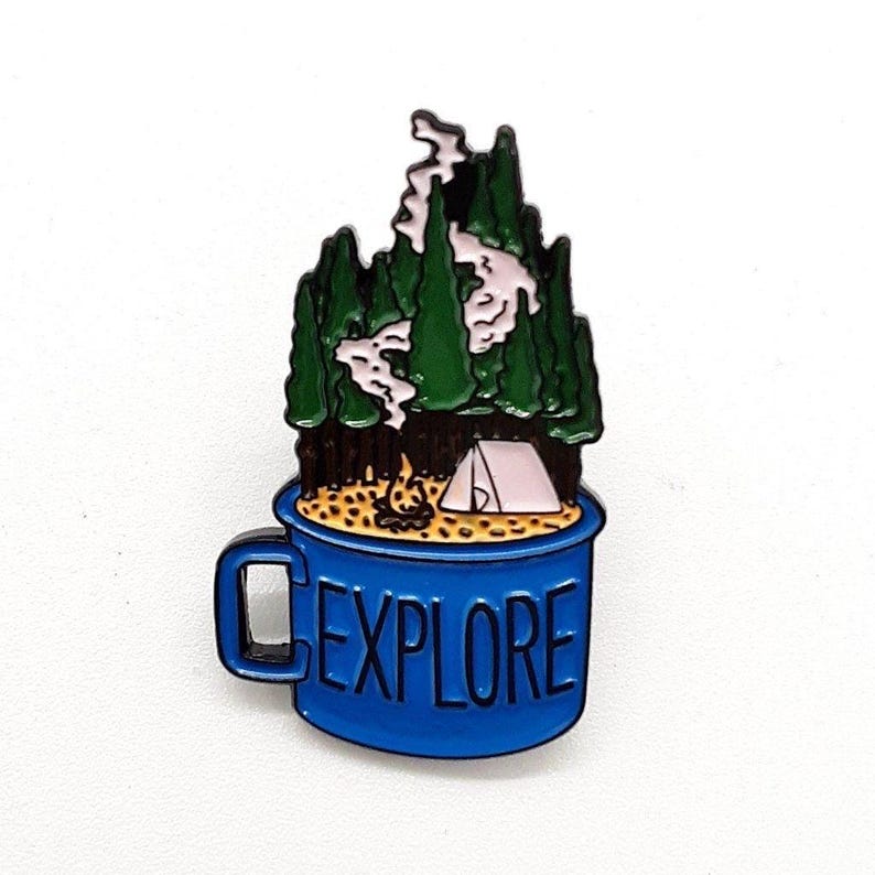 May include: A blue enamel pin shaped like a mug with the word "EXPLORE" on the side. The mug contains a camping scene with a tent, campfire, and trees. The trees are green, and the smoke is white.