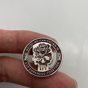 May include: A circular metal pin featuring a dark purple and silver design. The center depicts a skull topped with a rose. The outer border contains the text MAYBE WE CAN DROWN IN OUR DARKNESS TOGETHER with the letters BNA on the left and right sides.