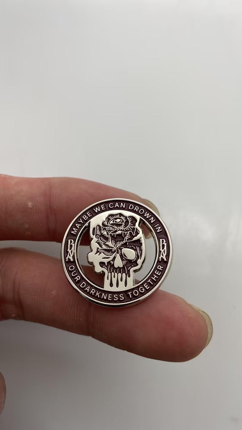 May include: A circular metal pin featuring a dark purple and silver design. The center depicts a skull topped with a rose. The outer border contains the text MAYBE WE CAN DROWN IN OUR DARKNESS TOGETHER with the letters BNA on the left and right sides.