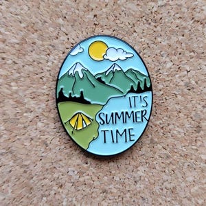 May include: Enamel pin with a camping theme. The oval pin features a blue sky with a yellow sun and white clouds, green mountains, and a yellow tent. The text "IT'S SUMMER TIME" is written on the pin.