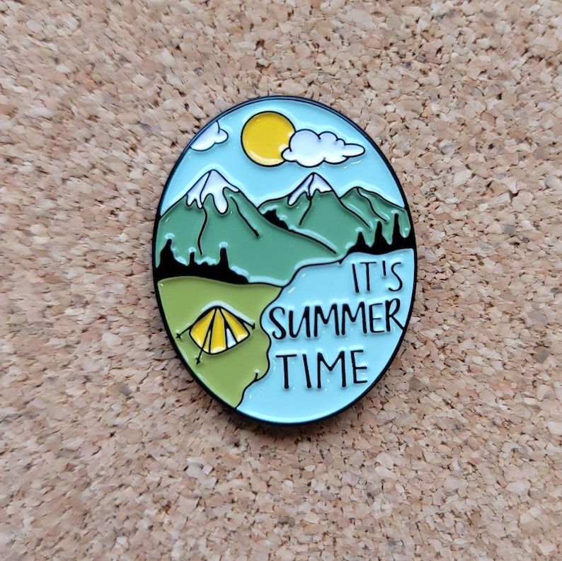May include: Enamel pin with a camping theme. The oval pin features a blue sky with a yellow sun and white clouds, green mountains, and a yellow tent. The text "IT'S SUMMER TIME" is written on the pin.