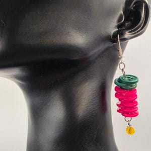 May include: A handmade earring featuring a stack of vibrant pink buttons topped with green buttons. A small yellow bead dangles at the bottom. The earring is suspended from a silver-colored fish hook.