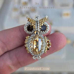 Pode incluir: Broche de coruja artesanal feito com contas douradas, prateadas e castanhas, com um grande cristal central âmbar. O texto "By Alexander Monneret" e "Planbead.com" está incluído.