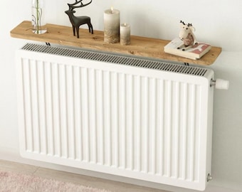 May include: A white panel radiator with vertical lines is fixed to a wall. Above, a wooden shelf displays a black deer ornament, candles, and a small book. A vase with a green plant is also on the shelf.