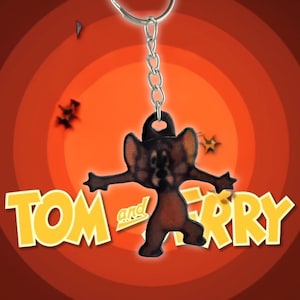 May include: A Tom and Jerry keychain featuring Jerry Mouse with outstretched arms. The keychain is brown and black, hanging from a silver chain. The background is a retro orange and red design with the words "TOM and JERRY" in yellow.