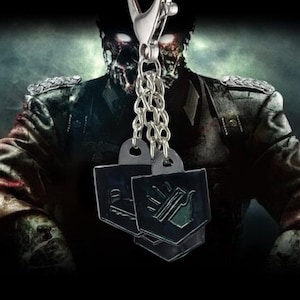 May include: A silver keychain with two black metal charms. Each charm has a unique symbol in green and white. The background features a blurred image of a zombie in a military uniform.