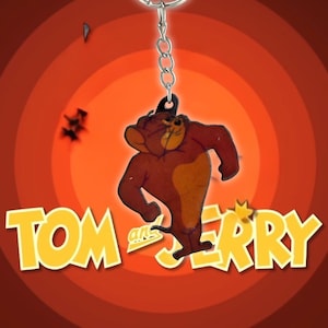 May include: A Tom and Jerry keychain featuring Jerry Mouse in a running pose. The keychain has a silver chain and a cartoon image of Jerry in brown and tan colors. The background is a red and orange gradient with the words "Tom and Jerry" in yellow.
