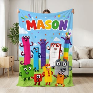 May include: A light blue fleece blanket featuring the name MASON in colorful block letters at the top. The design includes various Numberblocks characters in red, green, blue, purple, orange, and grey, set against a grassy field and sky.
