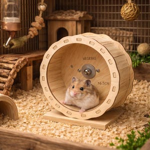 Peut inclure: Un hamster dans une cage en bois avec une roue en bois. La roue porte l'inscription "VEGEBIO" et "16,5 cm". La cage comprend également un biberon, une petite maison en bois et un pont en bois.
