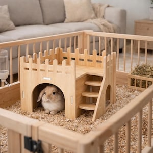 May include: A wooden rabbit enclosure with a castle-like structure, stairs, and a fenced perimeter. A small, light brown rabbit sits inside the structure. The enclosure is filled with wood shavings and includes a hay feeder and water bottle.