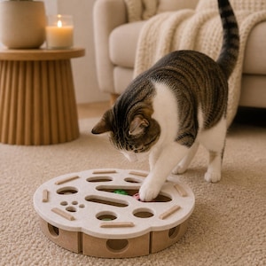 May include: A brown and white tabby cat interacting with a beige cat toy. The toy is round with multiple openings and contains colorful balls. The cat is reaching for a ball with its paw. A small wooden side table and a lit candle are in the background.