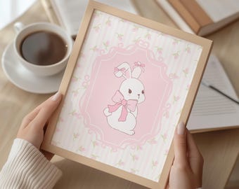 May include: A framed print with a cartoon bunny illustration. The bunny has a pink bow and is set against a pink background with a floral pattern. The frame is a light beige colour, and the print is held by a person.