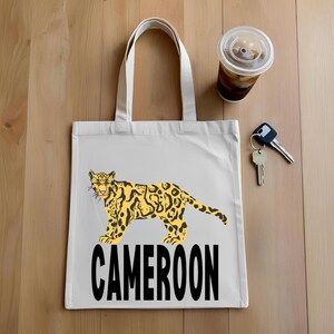 Puede incluir: Una bolsa de mano blanca con un diseño de leopardo amarillo y negro y la palabra "CAMEROON" en negro. La bolsa está sobre una superficie de madera, con una taza de café y llaves cerca. La bolsa tiene dos asas blancas.