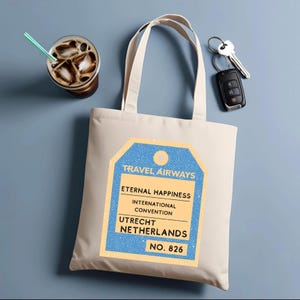 Puede incluir: Bolso tote color crema con un diseño de etiqueta de viaje azul y amarillo. La etiqueta dice "Travel Airways, Eternal Happiness, International Convention, Utrecht Netherlands, No. 826." Un vaso de café helado y llaves de coche están junto al bolso.