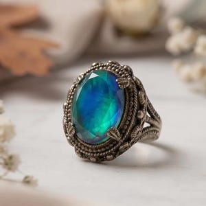Può includere: Un anello d'argento ovale con una grande pietra preziosa iridescente. La gemma mostra sfumature di blu e verde. L'anello ha un'incastonatura ornata e dettagliata con un design strutturato. L'anello è posto su uno sfondo morbido e sfocato.