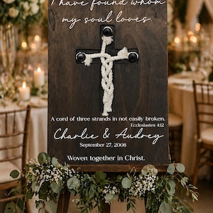 May include: A dark wooden wedding sign on an easel, featuring a rope cross and the inscription "I have found whom my soul loves." The sign also includes the names "Charlie & Audrey" and the date "September 27, 2008."