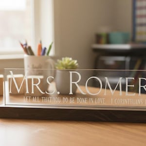 Puede incluir: Una placa de escritorio de acrílico transparente con el nombre "MRS. ROMERO" en letras blancas. La placa se asienta sobre una base de madera oscura. Debajo del nombre está el texto "LET ALL THAT YOU DO BE DONE IN LOVE 1 CORINTHIANS 16:14".