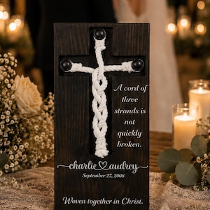 May include: A dark wood rectangular plaque featuring a white rope braided into a cross shape secured by three black studs. The text reads: A cord of three strands is not quickly broken. charlie & audrey September 27, 2008 Woven together in Christ.