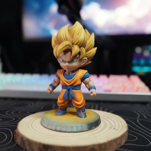 May include: A small, stylized figurine of a character with spiky blonde hair, wearing an orange martial arts uniform with a blue undershirt, blue wristbands, and a blue sash. The figure stands on a circular base with a yellow and grey finish.