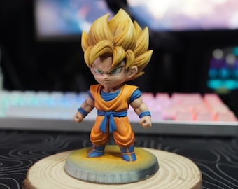 Può includere: Una piccola statuetta stilizzata di un personaggio con capelli biondi a punta, che indossa un'uniforme da arti marziali arancione con una maglietta blu, polsini blu e una fascia blu. La figura poggia su una base circolare con finitura.