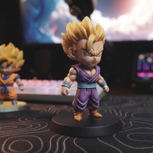 May include: A small, stylized figurine of a character with spiky blonde hair, wearing a purple martial arts uniform with a blue sash and tan boots, standing on a circular black base, with two other similar figures visible in the background.