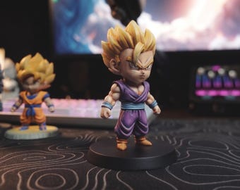 Può includere: Una piccola statuetta stilizzata di un personaggio con capelli biondi a punta, che indossa un'uniforme da arti marziali viola con una fascia blu e stivali color cuoio, in piedi su una base circolare nera, con altre due figure simili sullo sfondo.