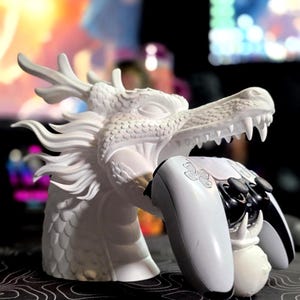May include: A white dragon head sculpture featuring detailed scales and flowing mane, designed as a stand to hold a white and black video game controller. The dragon's open mouth and clawed hand securely grip the controller in a horizontal position.
