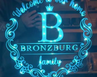 May include: A blue illuminated sign with the words "Welcome to our home" and "Bronzburg family". The design includes a large letter "B" with lights, a crown, and decorative swirls. The sign is designed for home decor.