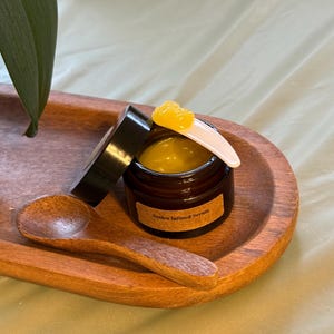 Puede incluir: Un pequeño frasco de vidrio marrón de suero con un aplicador blanco y una porción de producto amarillo en la parte superior. El frasco está sobre una bandeja de madera con una cuchara de madera. La etiqueta dice "Arnica Infused Serum".