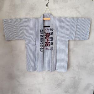以下が含まれることがあります： 水色と白のピンストライプ柄の法被。幅の広い袖と前開きのデザインが特徴です。前立ての両側には、濃い青色の日本語の漢字と円形の紋章が縦方向にプリントされています。