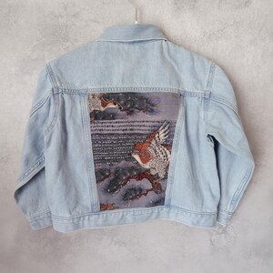 以下が含まれることがあります： 背中に鳥と枝のデザインが施された、明るいブルーのデニムジャケット。アートワークは、茶色、白、青の色合いが特徴です。ジャケットはシルバーのハンガーに掛けられています。