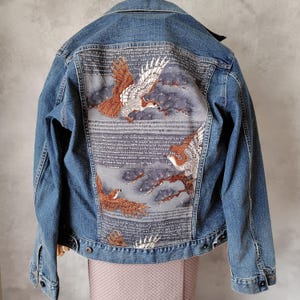以下が含まれることがあります： 背中に詳細な刺繍デザインが施された青いデニムジャケット。茶色と白の鳥が、灰色の雲と木々を背景に飛んでいるデザインです。ジャケットは、クラシックなボタンアップフロントです。