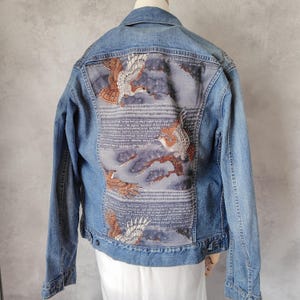 以下が含まれることがあります： 背中に刺繍のデザインが施された、薄い色のデニムジャケット。茶色、白、グレーの色合いの猛禽類が、様式化された雲と枝を背景に描かれています。ジャケットは、クラシックなボタンアップデザインです。