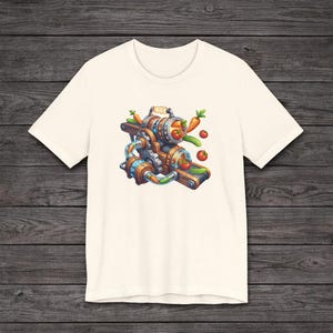 May include: Cream-colored t-shirt featuring a colorful illustration of a machine processing vegetables. The design includes carrots, tomatoes, and cucumbers. The machine has a steampunk aesthetic with gears and pipes.