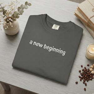May include: A charcoal grey t-shirt with the words "a new beginning" printed in white. The shirt is folded on a light grey surface. A small vase with flowers, a stack of books, a lit candle, and coffee beans are also visible.