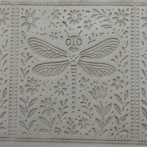 May include: A gray, rectangular stone tile with a carved dragonfly and floral design. The dragonfly is centered, with detailed wings and body. Flowers and leaves fill the surrounding space, with a decorative border.