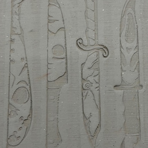 May include: A gray, textured surface with four knife designs carved into it. The knives vary in style, with details like serrated edges and decorative handles. The image is a close-up, highlighting the texture and detail of the carvings.