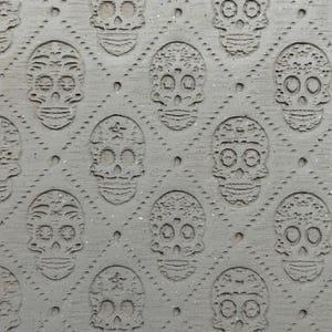 May include: Close-up of a gray surface with a repeating pattern of carved sugar skull designs. The skulls are detailed with eye sockets, nose cavities, and decorative elements. The background has a subtle speckled texture.