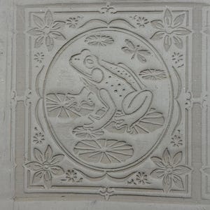 May include: A square, light gray tile with a carved design featuring a frog in a pond. The frog is surrounded by lily pads and dragonflies within a circular frame. Floral and geometric patterns border the central image.