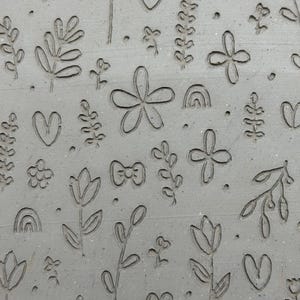May include: Close-up of a gray surface with an embossed floral pattern. The design includes flowers, hearts, bows, and leaves. The pattern is evenly spaced across the surface, creating a textured, decorative effect.